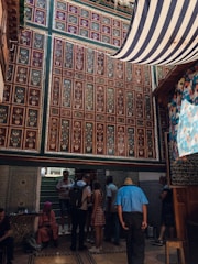 An intricately decorated interior features colorful geometric and floral patterns on the walls. A group of people, some seated and others standing, are gathered under a striped canopy. The setting appears to be lively yet relaxed.