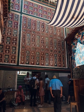 An intricately decorated interior features colorful geometric and floral patterns on the walls. A group of people, some seated and others standing, are gathered under a striped canopy. The setting appears to be lively yet relaxed.