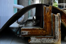 A small cat is resting within a makeshift shelter formed by large pieces of rusted metal and wood. The cat peeks out from the shadows, surrounded by industrial debris, under a curved sheet that provides cover.