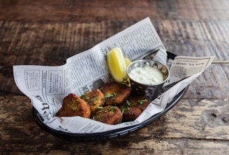 A basket lined with newspaper contains several crispy brown fritters garnished with herbs. Accompanying the fritters is a slice of lemon and a small metal cup filled with creamy dipping sauce. The basket is placed on a rustic wooden surface.