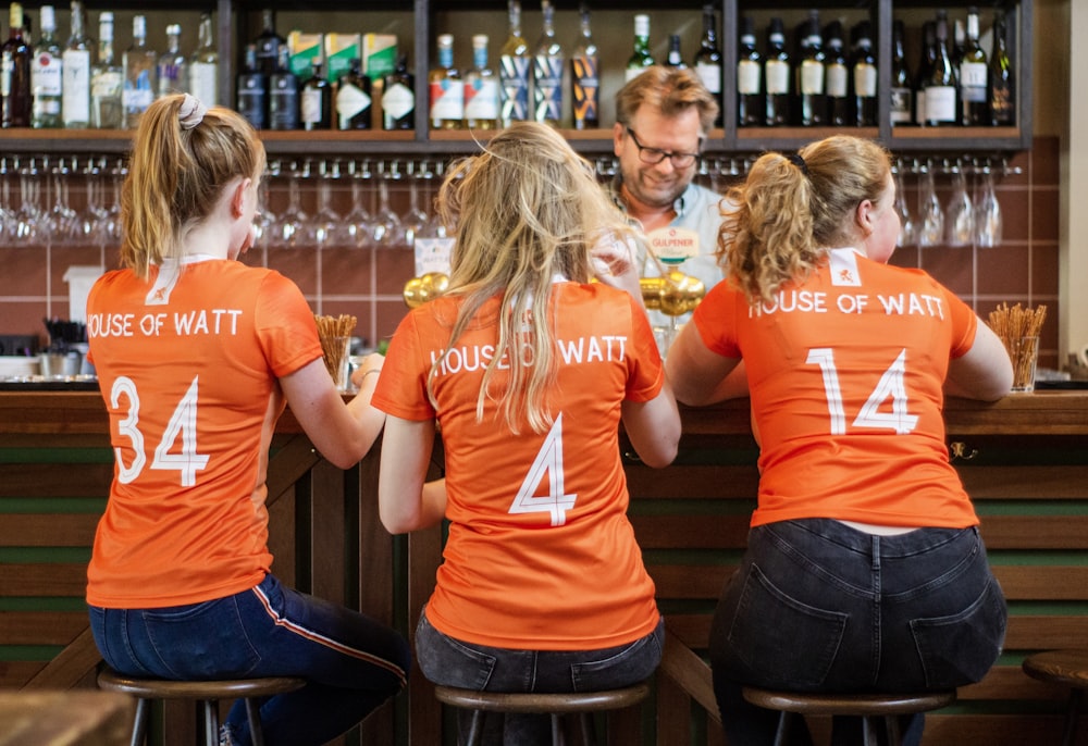 Tres mujeres con camisetas naranjas sentadas en taburetes de bar