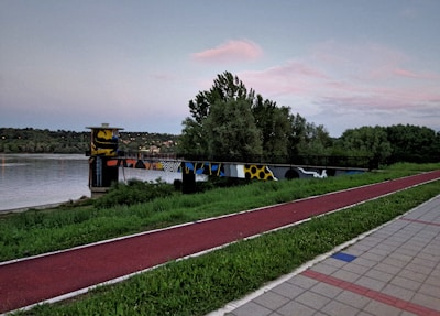 A vibrant walkway by a calm river, featuring a colorful structure with artistic graffiti. Lush green trees and bushes surround the area, set against a serene evening sky with hints of pink and blue. A red pathway runs parallel to the river, and part of it is paved.