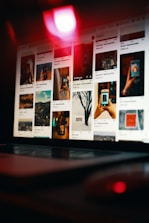 A laptop is displaying a Pinterest-like webpage, showing a grid of various images, including landscapes, close-up shots of hands holding a phone, and artistic photographs. The scene is dimly lit with a prominent red light reflecting off the screen, creating a moody ambiance.