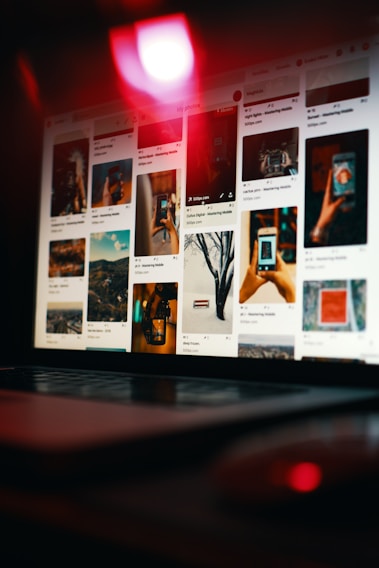 A laptop is displaying a Pinterest-like webpage, showing a grid of various images, including landscapes, close-up shots of hands holding a phone, and artistic photographs. The scene is dimly lit with a prominent red light reflecting off the screen, creating a moody ambiance.