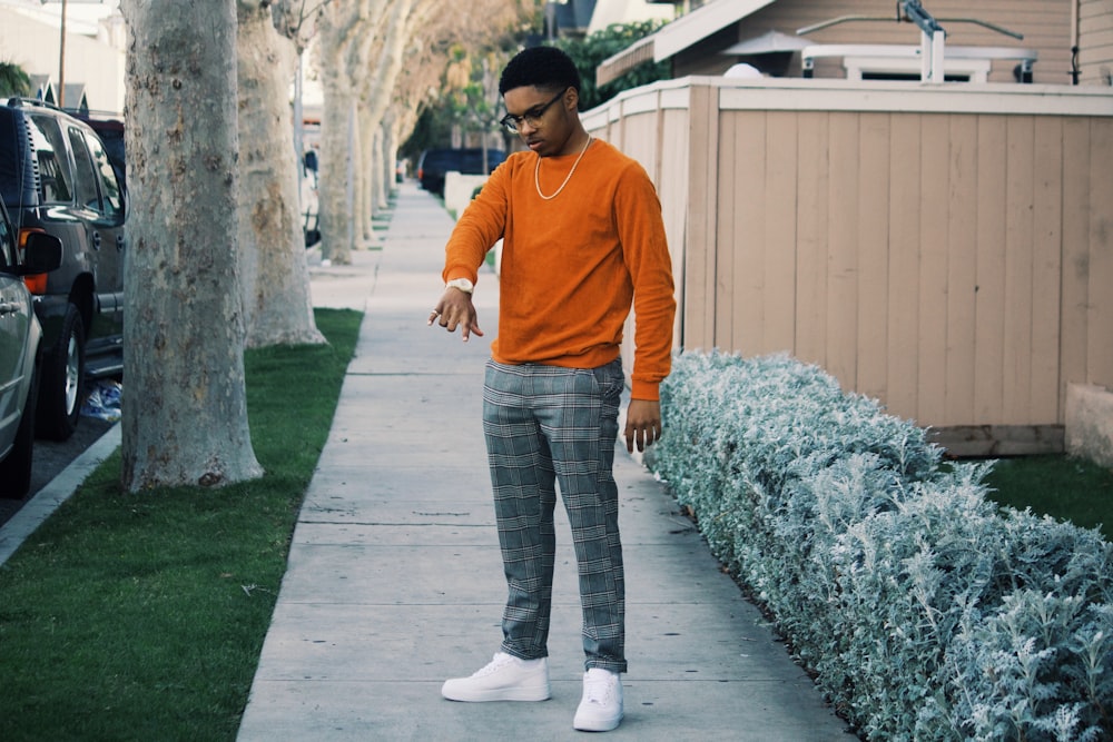 man in eyeglasses, orange top, and gray pants standing near plant