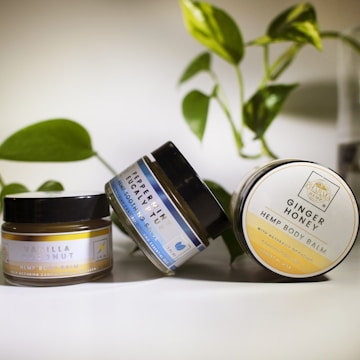 Three jars of hemp body balm with different scents, including Vanilla Coconut, Peppermint Eucalyptus, and Ginger Honey, are positioned on a surface with green leaves in the background. The jars are predominantly black with varying label designs indicating their respective scents.