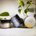 Three jars of hemp body balm with different scents, including Vanilla Coconut, Peppermint Eucalyptus, and Ginger Honey, are positioned on a surface with green leaves in the background. The jars are predominantly black with varying label designs indicating their respective scents.