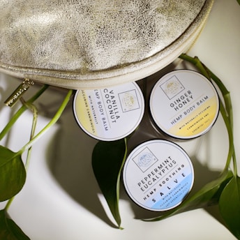 Three round tins of hemp body balm are arranged next to a textured metallic bag and some green leaves. The tins are labeled with different scents: Vanilla Coconut, Ginger Honey, and Peppermint Eucalyptus.