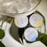 Three round tins of hemp body balm are arranged next to a textured metallic bag and some green leaves. The tins are labeled with different scents: Vanilla Coconut, Ginger Honey, and Peppermint Eucalyptus.