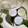 Three round tins of hemp body balm are arranged next to a textured metallic bag and some green leaves. The tins are labeled with different scents: Vanilla Coconut, Ginger Honey, and Peppermint Eucalyptus.