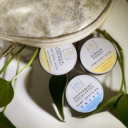 Three round tins of hemp body balm are arranged next to a textured metallic bag and some green leaves. The tins are labeled with different scents: Vanilla Coconut, Ginger Honey, and Peppermint Eucalyptus.