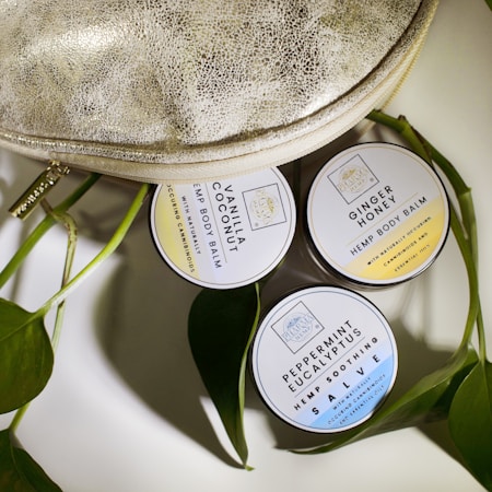 Three round tins of hemp body balm are arranged next to a textured metallic bag and some green leaves. The tins are labeled with different scents: Vanilla Coconut, Ginger Honey, and Peppermint Eucalyptus.