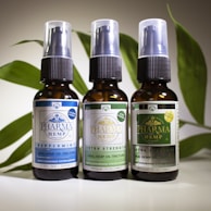 Three brown glass bottles with spray nozzles stand in a row, each labeled with various details about oral hemp oil tinctures. The labels have different colors: blue, light green, and dark green, indicating variations such as 'Peppermint,' 'Extra Strength,' and possibly another formula. Behind the bottles is a backdrop of green leaves, adding a natural element to the composition.