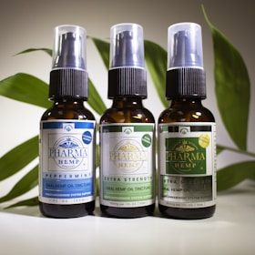 Three brown glass bottles with spray nozzles stand in a row, each labeled with various details about oral hemp oil tinctures. The labels have different colors: blue, light green, and dark green, indicating variations such as 'Peppermint,' 'Extra Strength,' and possibly another formula. Behind the bottles is a backdrop of green leaves, adding a natural element to the composition.