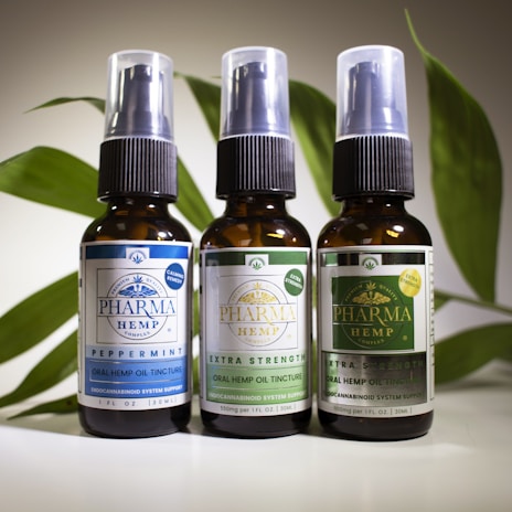 Three brown glass bottles with spray nozzles stand in a row, each labeled with various details about oral hemp oil tinctures. The labels have different colors: blue, light green, and dark green, indicating variations such as 'Peppermint,' 'Extra Strength,' and possibly another formula. Behind the bottles is a backdrop of green leaves, adding a natural element to the composition.