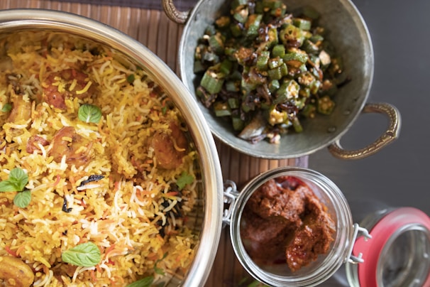 A meal featuring a large bowl of colorful rice dish with herbs, a side dish of chopped green vegetables seasoned in a metal container, and a jar of spicy-looking red condiment. The rice is garnished with fresh mint leaves, and the vegetable dish includes sliced okra.