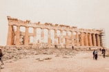 An ancient Greek temple with tall columns, partially in ruins, surrounded by scaffolding and a rocky terrain. Several people are walking and standing around, taking photos.