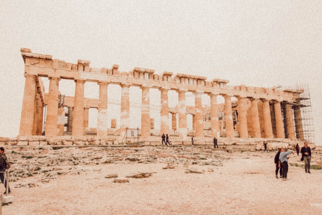 An ancient Greek temple with tall columns, partially in ruins, surrounded by scaffolding and a rocky terrain. Several people are walking and standing around, taking photos.