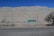 A semi-arid landscape with a road in the foreground and a directional road sign indicating 'Chuquicamata'. The background features large, barren hills or rock formations under a clear blue sky. Sparse vegetation with a few leafless trees and some power lines or pylons are visible.