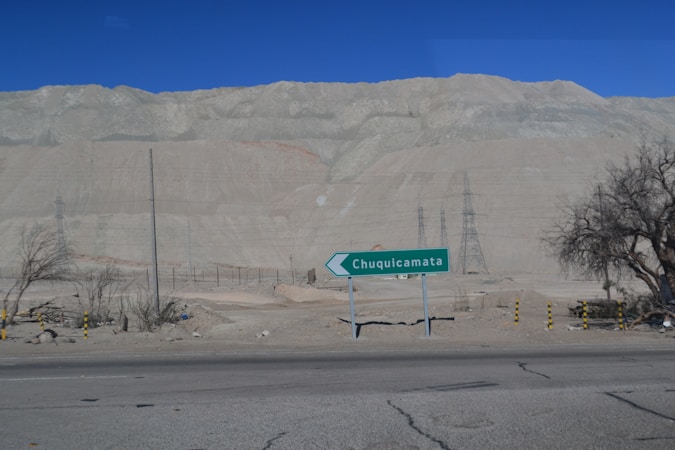 A semi-arid landscape with a road in the foreground and a directional road sign indicating 'Chuquicamata'. The background features large, barren hills or rock formations under a clear blue sky. Sparse vegetation with a few leafless trees and some power lines or pylons are visible.