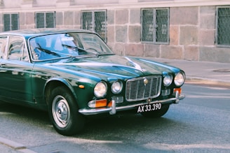 A classic vintage car, painted in a glossy dark green shade, is parked on a paved street against a stone building facade. The car's design features rounded headlights, chrome accents, and a license plate displaying 'AX 33.390'. The windows reflect the sky, adding a slightly nostalgic atmosphere to the scene.