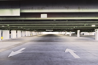 A spacious, empty parking garage with concrete pillars and ceiling. The floor has arrows painted on it indicating directions. The lighting is from overhead fixtures, providing a slightly dim and industrial atmosphere. A few cars are visible parked along the sides towards the back.