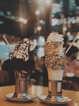 Two elaborately decorated milkshakes sit on a table. The left milkshake is adorned with whipped cream, chocolate sauce, and chocolate sprinkles, featuring a wide straw and a metal cup holder. The right milkshake is covered with colorful sprinkles and whipped cream, also with a straw and metal holder.