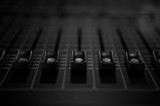 A close-up view of several audio mixing console faders arranged in a row. The faders are black with a metallic marker on each one. The background appears blurred, giving the image a shallow depth of field.
