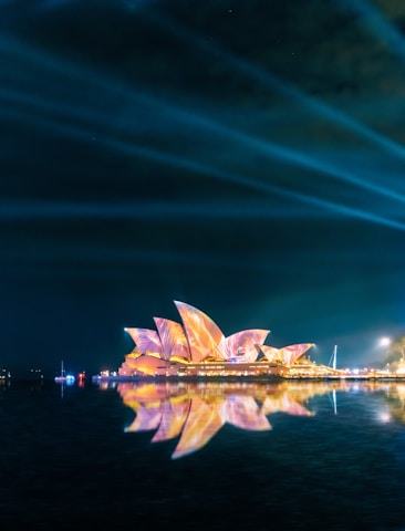 A vibrant night scene featuring the iconic Sydney Opera House illuminated with colorful lights, reflecting in the calm waters below. The sky is dark, with beams of light creating dynamic patterns.