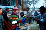 A street scene with a person in a colorful clown costume engaging with a street vendor. Surrounding them are various onlookers, including a man with a mustache and a girl in a green dress. The background reveals a busy street with a bus, palm trees, and other passersby. The vendor appears to be selling snacks displayed in front of him.