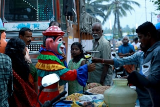A street scene with a person in a colorful clown costume engaging with a street vendor. Surrounding them are various onlookers, including a man with a mustache and a girl in a green dress. The background reveals a busy street with a bus, palm trees, and other passersby. The vendor appears to be selling snacks displayed in front of him.