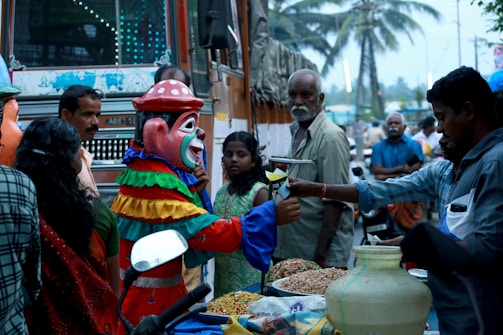 A street scene with a person in a colorful clown costume engaging with a street vendor. Surrounding them are various onlookers, including a man with a mustache and a girl in a green dress. The background reveals a busy street with a bus, palm trees, and other passersby. The vendor appears to be selling snacks displayed in front of him.