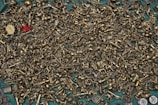 A wide array of small, intricate brass figurines and metallic objects are scattered across a teal surface. The assortment includes various miniature figures like animals, keys, and abstract shapes, with a prominent red object standing out amidst the monotone brass items.