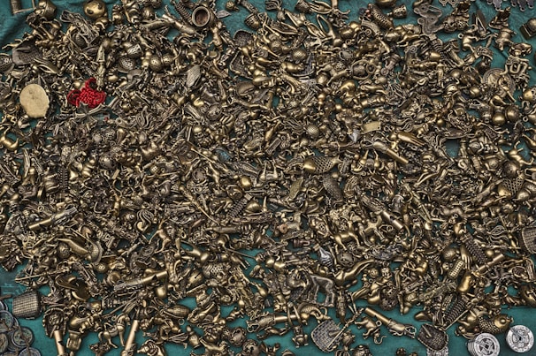 A wide array of small, intricate brass figurines and metallic objects are scattered across a teal surface. The assortment includes various miniature figures like animals, keys, and abstract shapes, with a prominent red object standing out amidst the monotone brass items.