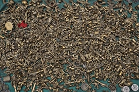 A wide array of small, intricate brass figurines and metallic objects are scattered across a teal surface. The assortment includes various miniature figures like animals, keys, and abstract shapes, with a prominent red object standing out amidst the monotone brass items.