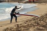 A person in a wetsuit is carrying a long red and white paddleboard up a sandy beach. The beach has gentle waves rolling in from a blue ocean. In the background, there are a few more beachgoers and orange buoys scattered along the shoreline.