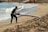 A person in a wetsuit is carrying a long red and white paddleboard up a sandy beach. The beach has gentle waves rolling in from a blue ocean. In the background, there are a few more beachgoers and orange buoys scattered along the shoreline.