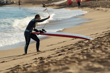 A person in a wetsuit is carrying a long red and white paddleboard up a sandy beach. The beach has gentle waves rolling in from a blue ocean. In the background, there are a few more beachgoers and orange buoys scattered along the shoreline.