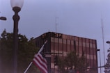 A tall, modern glass building with digital temperature displays reading 77°F and 71°F. In the foreground, an American flag is partially visible near a streetlamp. Surrounding the building are trees and various street signs, with a cloudy sky above.
