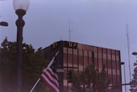 A tall, modern glass building with digital temperature displays reading 77°F and 71°F. In the foreground, an American flag is partially visible near a streetlamp. Surrounding the building are trees and various street signs, with a cloudy sky above.