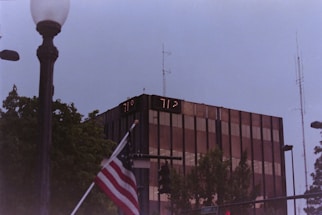 A tall, modern glass building with digital temperature displays reading 77°F and 71°F. In the foreground, an American flag is partially visible near a streetlamp. Surrounding the building are trees and various street signs, with a cloudy sky above.