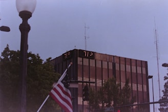 A tall, modern glass building with digital temperature displays reading 77°F and 71°F. In the foreground, an American flag is partially visible near a streetlamp. Surrounding the building are trees and various street signs, with a cloudy sky above.