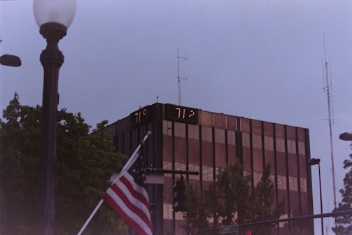 A tall, modern glass building with digital temperature displays reading 77°F and 71°F. In the foreground, an American flag is partially visible near a streetlamp. Surrounding the building are trees and various street signs, with a cloudy sky above.