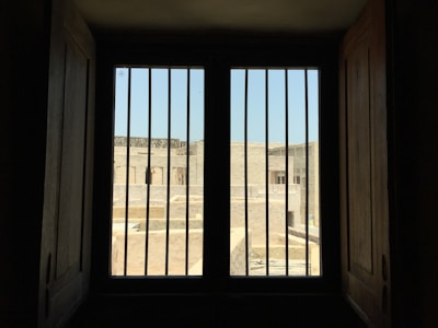 A barred window provides a view of a sunlit outdoor area with stone buildings. The scene is framed by dark wooden shutters, creating a contrast between the dimly lit interior and the bright exterior.