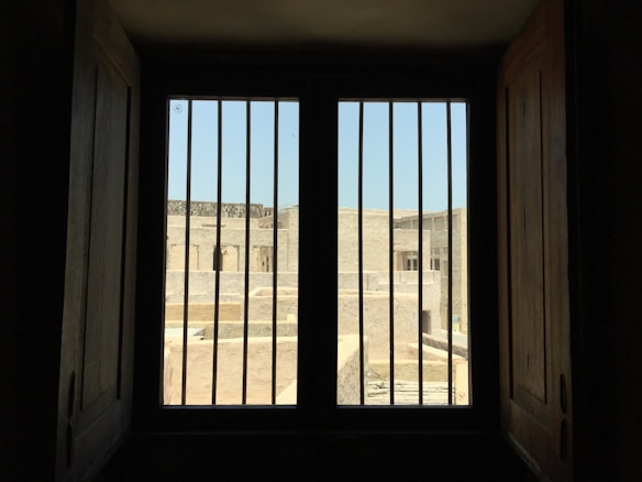 A barred window provides a view of a sunlit outdoor area with stone buildings. The scene is framed by dark wooden shutters, creating a contrast between the dimly lit interior and the bright exterior.