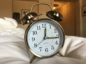 A classic brass twin bell alarm clock with white clock face and black numerals is placed on a soft, white surface, likely a bed. In the background, a blurred interior with warm lighting includes a framed picture and a decorative object on the wall.
