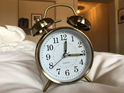 A classic brass twin bell alarm clock with white clock face and black numerals is placed on a soft, white surface, likely a bed. In the background, a blurred interior with warm lighting includes a framed picture and a decorative object on the wall.