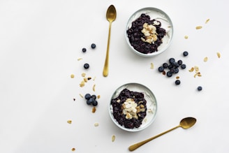 Two bowls filled with creamy yogurt are topped with blueberry compote and almond flakes, surrounded by scattered blueberries and almond slices on a clean, white surface. Two golden spoons are positioned nearby.