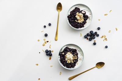Two bowls filled with creamy yogurt are topped with blueberry compote and almond flakes, surrounded by scattered blueberries and almond slices on a clean, white surface. Two golden spoons are positioned nearby.