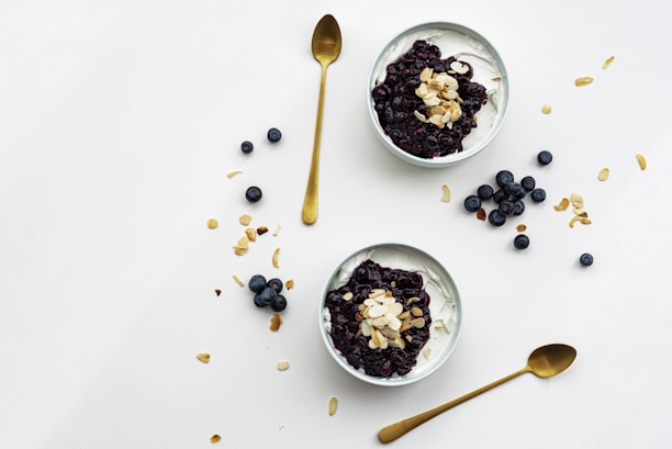 Two bowls filled with creamy yogurt are topped with blueberry compote and almond flakes, surrounded by scattered blueberries and almond slices on a clean, white surface. Two golden spoons are positioned nearby.